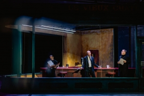 Mroczne wnętrze małego baru oglądanego jak przez szybę: czerwony długi blat, kilka czerwonych stołków, ciepłe żółto-rdzawe ściany, nad nimi zimna świetlówka. Trzech mężczyzn w czerni — dwaj po bokach trzymają kartki, środkowy w pasiastym garniturze opiera się o blat — między szklankami wody stoją termos i kopczyk cytryn.