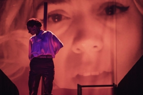 Scena w czerwonym świetle. Po lewej sylwetka kobiety w luźnej białej bluzce i ciemnych spodniach, z opuszczoną głową, oparta o pionową belkę. W tle ogromna projekcja twarzy — zbliżenie oczu, nosa i ust.