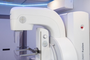 Jasne, sterylne wnętrze mammobusu: duży biały aparat do mammografii z ramieniem i przezroczystymi płytkami kompresyjnymi; nad nim chłodne, niebieskie oświetlenie LED z geometrycznym sufitem, w tle drzwi i klimatyzator.