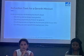 Sala szkoleniowa w półmroku: na ekranie rzutnika slajd. Przy stole siedzą dwie kobiety z laptopami, wokół porozkładane papiery i butelki. Atmosfera skupionej pracy warsztatowej.