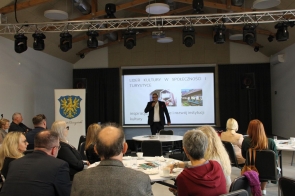 Sala konferencyjna z publicznością przy okrągłych stolikach; na scenie mężczyzna w garniturze mówi do mikrofonu przed ekranem z prezentacją „Lider kultury w społeczności i turystyce”. Po lewej roll-up z herbem Powiatu Cieszyńskiego, nad sceną zawieszone oświetlenie.