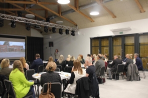 Sala konferencyjna z wysokim, drewnianym stropem; przy ścianie po lewej duży ekran z projekcją budynku w remoncie. Przed ekranem przemawia kobieta z mikrofonem, a kilkadziesiąt osób siedzi przy okrągłych stolikach, słuchając prezentacji.