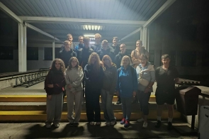 Pod zadaszeniem, na niskich schodach z żółtymi krawędziami, stoi kilkunastoosobowa grupa, głównie młodzież oraz dwie kobiety z przodu, po prawej widać kosz i stojącą walizkę.