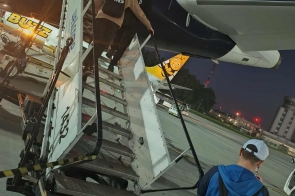 Na płycie lotniska grupa młodych z plecakami wchodzi po metalowych schodach do samolotu, z przodu chłopak w niebieskiej bluzie i czapce z plecakiem Adidas, w kadrze widoczne skrzydło i ogon z logo żurawia.