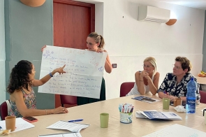 Cztery kobiety w jasnej sali siedzą przy stole podczas warsztatów. Jedna stoi i trzyma duży plakat z napisem „TEACHER”, druga wskazuje na treść plakatu; pozostałe dwie uważnie słuchają. Na stole leżą notatki, markery i kubki, w tle klimatyzator i proste wyposażenie.