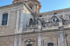 Kamienna fasada kościoła z monumentalną kopułą w żółto-zielone zygzaki, ozdobny balkon z balustradą i masywny portal; obok stoją stylowe latarnie, nad wszystkim czyste, błękitne niebo.