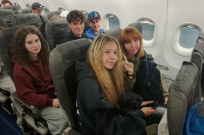 W samolocie, przy oknach w rzędzie szarych foteli, siedzi pięcioro nastolatków zwróconych do aparatu: najbliżej dziewczyna z długimi jasnymi falami i telefonem w dłoni, obok rudowłosa z plecakiem, za nimi uśmiechnięci chłopcy (jeden w czapce z lustrzanymi okularami na daszku), a po lewej dziewczyna o ciemnych kręconych włosach w bordowej bluzie.