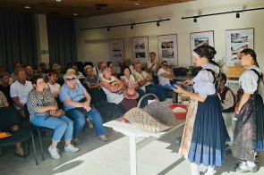 ChatGPT powiedział:  Sala spotkań wypełniona publicznością siedzącą w półkolu. Z przodu trzy kobiety w strojach ludowych prowadzą pokaz przy białym stole, na którym leży wiklinowy kosz i rekwizyty. Promienie słońca wpadają przez okno, na ścianach wiszą plansze, a nad głowami widać drewniany sufit z projektorem.