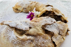 Złociste, cienkie ciasto zawijane, hojnie oprószone cukrem pudrem; w przekroju widać wilgotne, owocowe nadzienie z rodzynkami. Podane na białej, koronkowej serwetce, obok drobne kwiatki jako dekoracja.