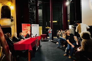 Panel dyskusyjny na scenie teatru, przy długim stole z bordowym obrusem siedzi kilka osób z mikrofonami, obok stoją roll-upy festiwalowe. Po prawej stronie niewielka publiczność – część fotografuje, kamerzysta nagrywa wydarzenie. Wnętrze bogato zdobione, z balkonem i złoceniami po lewej, wysoko wiszą bordowe kotary.