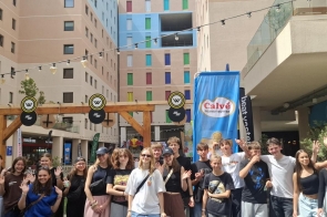 Rozbawiona grupa młodzieży pozuje na dziedzińcu między budynkami z kolorowymi okiennicami; wielu macha do aparatu. W tle czarne i niebieskie flagi reklamowe, drewniana brama i girlandy lampe.