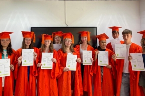 Uśmiechnięci nastolatkowie ustawieni w rzędzie, wszyscy w jaskrawo pomarańczowych togach i biretach z chwostami. Każdy trzyma przed sobą certyfikat ukończenia, stoją w jasnym pomieszczeniu