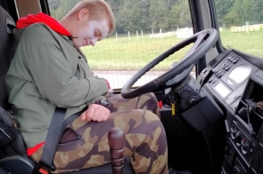 Młody mężczyzna z pobladłą twarzą siedzi zapięty pasem w kabinie ciężarówki, głowa opada na pierś jakby nieprzytomny, wokół widać kierownicę i przyrządy, za szybą zielona łąka i las.