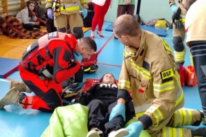 Ratownicy i strażacy udzielają pomocy leżącej osobie w szkolnej sali gimnastycznej, głowa na czerwonej desce, nogi oparte o zielony puf.