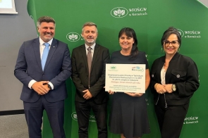 Czworo dorosłych w eleganckich strojach stoi przy zielonej ściance z logotypem „WFOŚiGW w Katowicach”. Kobieta pośrodku trzyma dużą tablicę informującą o dofinansowaniu „Ekopracowni pod chmurką”. Wszyscy uśmiechnięci.