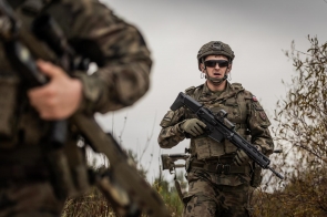 Żołnierz w hełmie i mundurze polowym stoi z karabinem w rękach na otwartym terenie. Na pierwszym planie widoczny fragment sylwetki innego żołnierza z bronią.
