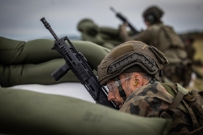 Żołnierz w hełmie i mundurze polowym celuje z karabinu zza umocnień z worków z piaskiem. W tle widoczny drugi żołnierz w podobnej pozycji.