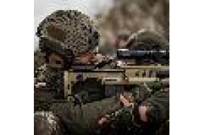 Żołnierz w hełmie i mundurze celuje z karabinu wyposażonego w optykę. Broń oparta na ramieniu, widoczny fragment drugiego żołnierza w tle.