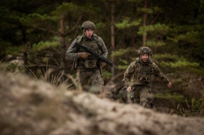 Dwóch żołnierzy w mundurach polowych i hełmach porusza się w lesie z karabinami w rękach. Pierwszy żołnierz idzie z przodu, drugi nieco z tyłu, w tle widoczne drzewa.