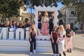 Grupa nastolatek pozuje przy dużej, białej instalacji z ozdobną ramką, część stoi w środku ramy. Słoneczny plac, wokół wysokie drzewa i jasne, zabytkowe budynki