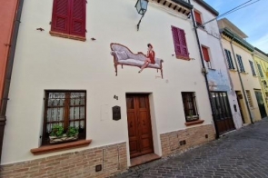 Fasada wąskiego domu z numerem 41, kremowe ściany z muralem kobiety na leżance, bordowe okiennice i drewniane drzwi. Pod oknami kwiaty, przed domem wąska, brukowana uliczka.