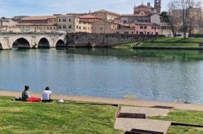 Zielona skarpa nad rzeką, na trawie siedzą dwie osoby z kocem, patrzą na wodę. Po lewej kamienny most z kilkoma półkolistymi przęsłami. Po drugiej stronie rzeki pastelowe kamienice i wieża kościoła.