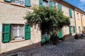 Ceglana kamienica z zielonymi okiennicami; nad kutą, łukową bramą zwiesza się gęste pnącze. Wąska brukowana uliczka, z boku oparte dwa rower.