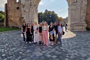 Grupa kilkunastu młodych kobiet pozuje pod ogromnym, jasnym kamiennym łukiem w słoneczny dzień. Stoją na brukowanym placu, część ma jasne, letnie stroje, część ciemniejsze, wszyscy uśmiechnięci. W tle widać fragment starego muru i zielone drzewa oświetlone popołudniowym słońcem.