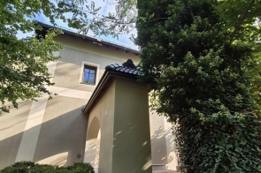 Fragment budynku z jasną elewacją i ciemnym dachem otoczony roślinnością. Na ścianach widoczny jest bluszcz, a w tle znajdują się drzewa i niebo.