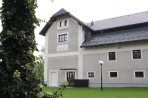 Budynek o jasnej elewacji z dwuspadowym dachem pokrytym ciemnymi dachówkami. Na parterze znajduje się wejście z brązowymi drzwiami i kilka prostokątnych okien. Obok stoi latarnia, a w tle widoczne są drzewa i trawnik.