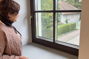 Janina Żagan Starosta Cieszyński stoi przy oknie i patrzy na zewnątrz. Widać fragment parapetu, drewnianą ramę okienną oraz ogród i dom po drugiej stronie ulicy.  