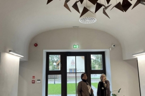 Dwie osoby stoją w holu przy wejściu z przeszklonymi drzwiami. Na suficie zawieszona jest dekoracja z ciemnych elementów, a po bokach widać oświetlenie ścienne i roślinę w doniczce.