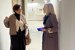 Dwie kobiety stoją w jasnym korytarzu i rozmawiają. Obie trzymają torby i są ubrane w płaszcze. W tle widać drzwi, ściany z oznaczeniami i sufit z oświetleniem.