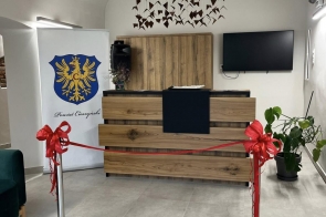 Nowoczesne wnętrze z drewnianą ladą recepcyjną i minimalistycznym wystrojem. Przed ladą rozciągnięta jest czerwona wstęga ozdobiona kokardami, przygotowana do przecięcia. Nad ladą wisi dekoracja z zawieszonych trójkątnych elementów, a obok znajduje się roślina doniczkowa i telewizor na ścianie. Po lewej stronie stoi roll-up z herbem i napisem „Powiat Cieszyński”.