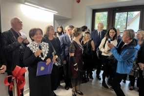 Grupa elegancko ubranych osób stoi w jasnym pomieszczeniu podczas uroczystego wydarzenia. Kobieta z mikrofonem w dłoni przemawia, uśmiechając się do zgromadzonych. Wokół niej znajdują się ludzie w formalnych strojach — niektórzy mają garnitury, inne panie stylizacje z perłami i ozdobami w stylu retro. W tle widać okno, a kilka osób fotografuje lub nagrywa wydarzenie telefonem.