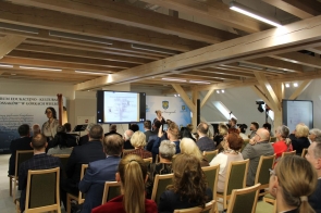 W sali z drewnianymi belkami na suficie trwa prezentacja dla licznie zgromadzonej publiczności. Kobieta przy mikrofonie stoi przed dużym ekranem, na którym wyświetlony jest schemat drzewa genealogicznego rodziny Kossaków. Uczestnicy siedzą w rzędach, uważnie słuchając i obserwując wystąpienie. W tle widoczne są banery z nazwą „Centrum Edukacyjno-Kulturalne U Kossaków w Górkach Wielkich” oraz herb powiatu.