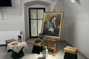 W jasnym pomieszczeniu stoi sztaluga z dużym obrazem przedstawiającym osobę pogrążoną w pisaniu. Obok znajdują się drewniane stołki, na jednym z nich siedzi lalka w niebieskim ubraniu. Na małym stoliku przykrytym koronkową serwetką leżą książki i zdjęcie, a na podłodze stoi biała latarenka. Całość oświetlona jest ciepłym światłem z lampy wiszącej pod sufitem.