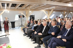 Kobieta w czerwonej sukni z ozdobnymi detalami występuje z mikrofonem przed publicznością w jasnej sali z drewnianymi belkami na suficie. Słuchacze siedzą w rzędach, elegancko ubrani w garnitury i formalne stroje, uśmiechają się i słuchają z zainteresowaniem. Na pierwszym planie widać kwiaty i fragment sceny, a w tle sprzęt nagłośnieniowy i stojaki oświetleniowe.