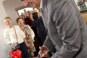 Elegancko ubrany mężczyzna w szarym garniturze przecina dużymi nożyczkami czerwoną wstęgę. Za nim stoi grupa osób obserwujących moment z uśmiechem, niektórzy robią zdjęcia telefonami. W tle widać kwiaty, elementy wystroju sali i biały baner z herbem.