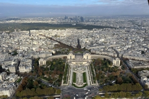 Panorama dużego miasta widziana z wysokości. W centralnej części rozciąga się rozległy plac z ogrodami i dwoma symetrycznymi budynkami w stylu klasycznym. Wokół widać gęsto zabudowane ulice i jasne kamienice, a w oddali nowoczesną dzielnicę z wieżowcami. Niebo jest częściowo zachmurzone, a światło słoneczne delikatnie oświetla miasto.