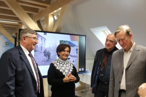 Cztery osoby od lewej: Jerzy Pilch, Janina Żagan, Franciszek i Michał Rosset stoją razem w jasnym pomieszczeniu i rozmawiają z uśmiechem. Kobieta w czarnej sukni z białym koronkowym kołnierzem znajduje się w centrum grupy, obok trzech elegancko ubranych mężczyzn w garniturach. W tle widać ekran z czarno-białym zdjęciem przedstawiającym dwie osoby przed starym budynkiem oraz drewniane belki konstrukcyjne poddasza.