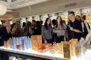 Grupa młodych osób znajduje się w eleganckim sklepie z perfumami. Wnętrze jest jasno oświetlone, z ozdobnym sufitem i półkami pełnymi kolorowych flakonów. Na pierwszym planie stoi kilka dziewczyn trzymających paski testowe do zapachów, a przed nimi leży stół z pomarańczowymi i niebieskimi opakowaniami perfum. W tle widać innych uczestników oglądających produkty.