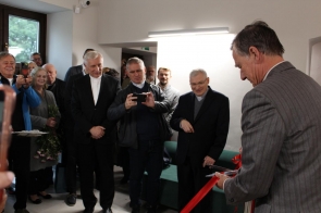 Mężczyzna w szarym garniturze przecina czerwoną wstęgę, a wokół niego stoi grupa uśmiechniętych osób obserwujących ten moment. Kilku mężczyzn w ciemnych garniturach, w tym duchowni w koloratkach, robi zdjęcia telefonami. W tle widać okno, zieloną kanapę i osoby trzymające kwiaty.