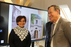 Janina Żagan w czarnej sukni z białym koronkowym kołnierzem stoi obok dwóch uśmiechniętych mężczyzn. Wszyscy rozmawiają i wyglądają na zadowolonych. W tle widać ekran z prezentacją pokazującą zdjęcia starego budynku i jego odnowionej fasady, a za nimi drewniane belki konstrukcyjne poddasza.