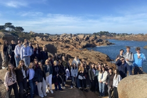 Grupa młodych osób stoi razem na skalistym wybrzeżu z dużymi różowymi głazami. W tle widać spokojne morze i linie brzegową z niskimi drzewami. Niebo jest bezchmurne i jasnoniebieskie, a słońce oświetla całą scenę. Ludzie są uśmiechnięci i ubrani w luźne, codzienne stroje.