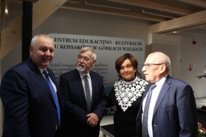 Cztery elegancko ubrane osoby stoją razem i rozmawiają w jasnym pomieszczeniu. Kobieta w czarnej sukni z białym koronkowym kołnierzem stoi pośrodku, otoczona przez trzech mężczyzn w garniturach i krawatach. W tle widać tablicę z napisem „Centrum Edukacyjno-Kulturalne U Kossaków w Górkach Wielkich” oraz fragment cytatu Zofii Kossak. Wszyscy uśmiechają się i wyglądają na zadowolonych.