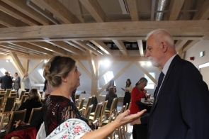 Kobieta w eleganckiej, ciemnej sukni ozdobionej cekinami rozmawia z mężczyzną w garniturze i krawacie. Stoją w jasnej sali z drewnianymi belkami i rzędami krzeseł, gdzie trwa przerwa w wydarzeniu. W tle kilka osób siedzi lub rozmawia przy stolikach, a jedna z kobiet pije kawę.