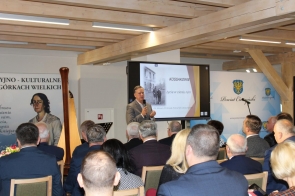 Mężczyzna w szarym garniturze stoi przed publicznością i przemawia, trzymając mikrofon. Za nim znajduje się ekran z prezentacją zatytułowaną „Kossakowie – życie w cieniu ruin” oraz logo powiatu na tle jasnoniebieskiej ścianki. W sali siedzi wiele osób w eleganckich strojach, słuchających wystąpienia. Po lewej stronie widać harfę i fragment obrazu przedstawiającego kobietę.