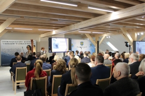 W przestronnej sali z drewnianymi belkami na suficie trwa uroczystość z udziałem licznej publiczności. Ludzie siedzą w rzędach, zwróceni w stronę mównicy i dużego ekranu z prezentacją zatytułowaną „Kossakowie – życie w cieniu ruin”. Na scenie przemawia kobieta, obok której stoi harfa. W tle widoczny jest baner z nazwą „Centrum Edukacyjno-Kulturalne U Kossaków w Górkach Wielkich” oraz logo powiatu.