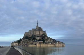 Duże kamienne opactwo wznosi się na skalistej wyspie otoczonej wodą. Budowla ma wysoką wieżę z iglicą, a u jej stóp rozciągają się mniejsze domy i mury obronne. Do wyspy prowadzi długa grobla, po której idą ludzie. Niebo jest lekko zachmurzone, a woda spokojna i odbija kolory nieba.